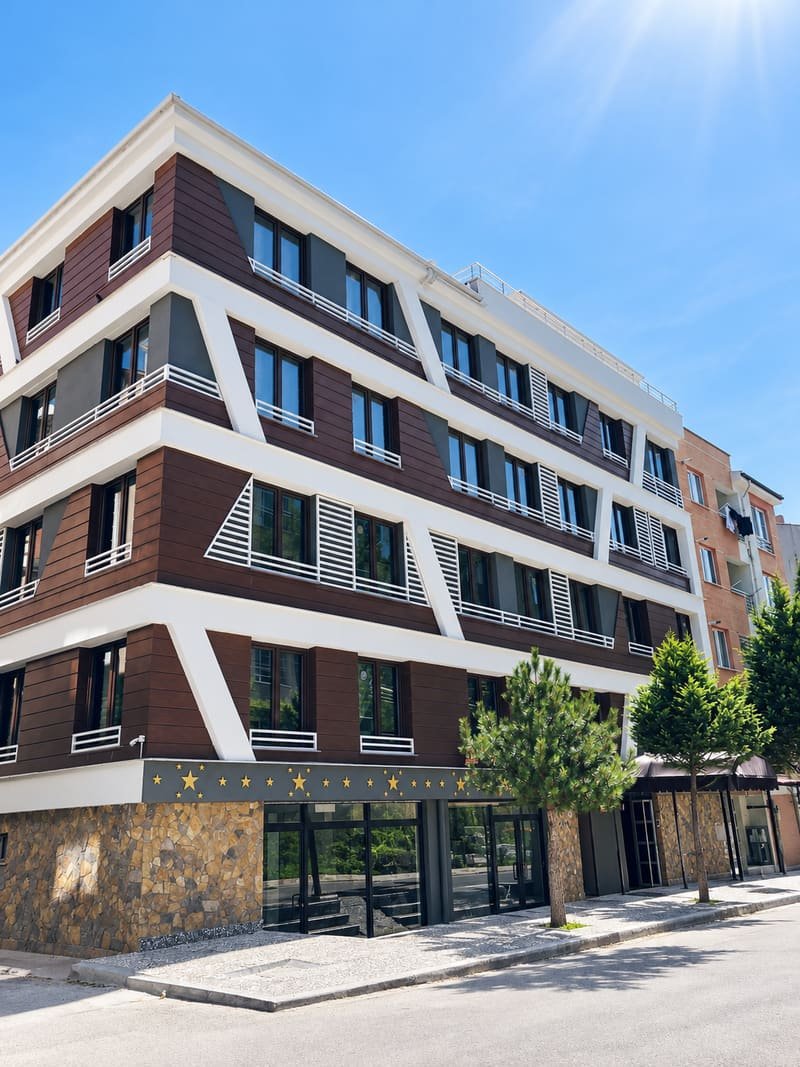 EsVia Suit Apart binasının köşeden görünümü: kahverengi kaplamalı, beyaz bantlı cephe, zemin katta taş kaplama ve cam giriş, önünde ağaçlar